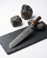 Couteau japonais type Santoku en acier damassé posé sur une plaque d’ardoise noire, manche en bois brun veiné, pierres sombres autour.
