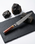 Couteau japonais type Petty en acier damassé posé sur une plaque d’ardoise noire, manche en bois brun veiné, pierres sombres en décor.