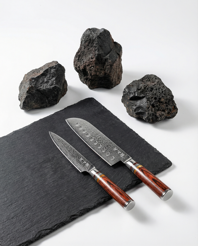 Deux couteaux japonais en acier damassé posés sur une plaque d’ardoise noire, manches en bois brun veiné, pierres volcaniques en arrière-plan.
