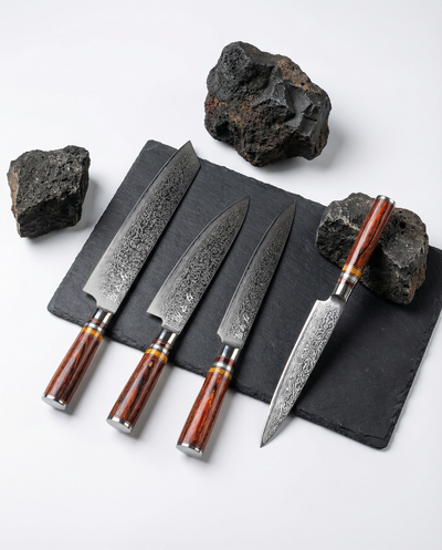 Quatre couteaux japonais en acier damassé posés sur une plaque d’ardoise noire, manches en bois brun veiné, pierres sombres autour.