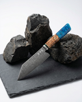 Couteau japonais Bunka en acier damassé posé sur ardoise noire, manche en bois naturel et résine bleue, pierres volcaniques en décor.