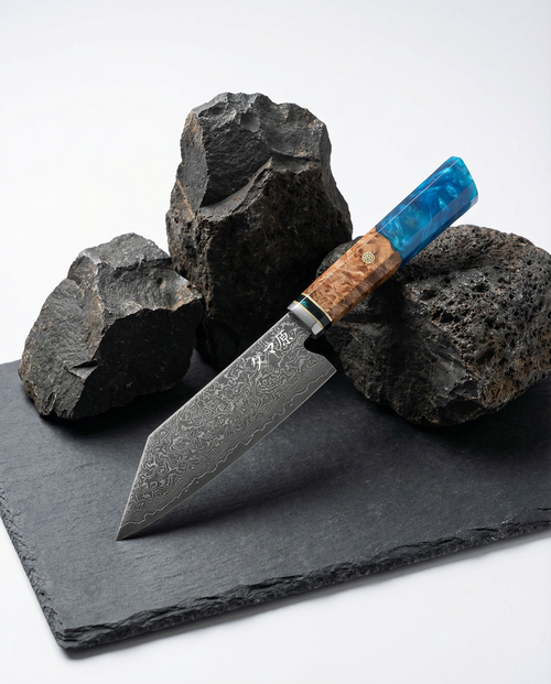 Couteau japonais Bunka en acier damassé posé sur ardoise noire, manche en bois naturel et résine bleue, pierres volcaniques en décor.