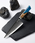 Couteau japonais Santoku en acier damassé posé sur ardoise noire, manche en bois naturel et résine bleue, pierres volcaniques autour.