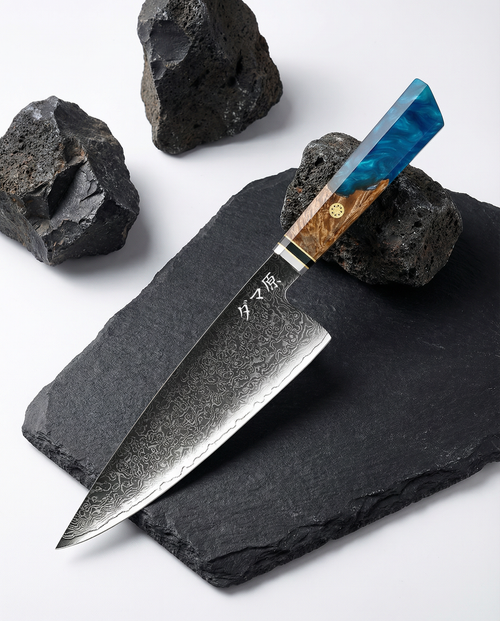 Couteau japonais Santoku en acier damassé posé sur ardoise noire, manche en bois naturel et résine bleue, pierres volcaniques autour.