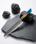 Couteau japonais Gyuto en acier damassé posé sur ardoise noire, manche en bois naturel et résine bleue, pierres volcaniques en décor.