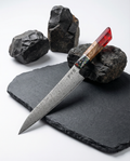 Couteau japonais damassé Gyuto, lame longue en acier Damas motif martelé, manche bois naturel avec insert multicolore et résine rouge, présenté sur ardoise noire avec pierres volcaniques.