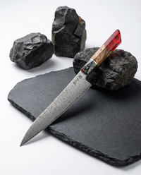 Couteau japonais damassé Gyuto, lame longue en acier Damas motif martelé, manche bois naturel avec insert multicolore et résine rouge, présenté sur ardoise noire avec pierres volcaniques.