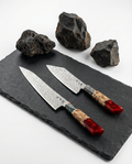 Couteaux japonais damassés duo, lames en acier Damas motif ondulé, manches bois naturel et résine rouge, présentés sur ardoise noire avec pierres volcaniques.