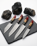 Set de quatre couteaux japonais damassés disposés sur ardoise noire, lames martelées aux motifs ondulés, manches en bois clair avec résine rouge translucide et insert multicolore, pierres volcaniques en arrière-plan.