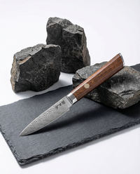 Couteau d’office japonais en acier damassé avec manche en bois brun, posé sur ardoise noire et pierres volcaniques.