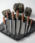 Set de 7 couteaux japonais en acier damassé, manches bois naturel, présentés sur ardoise noire avec pierres volcaniques et romarin.