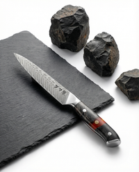Couteau japonais petty en acier damas, lame fine gravée, manche résine noir et ambre, posé sur ardoise avec pierres volcaniques.