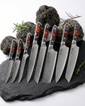Set de 8 couteaux japonais en acier damassé avec manches noir et rouge, disposés sur ardoise noire avec pierres volcaniques et branches de romarin.