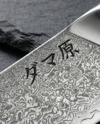 Gros plan sur lame en acier damassé d’un couteau japonais, motifs ondulés contrastés et gravure de caractères noirs.