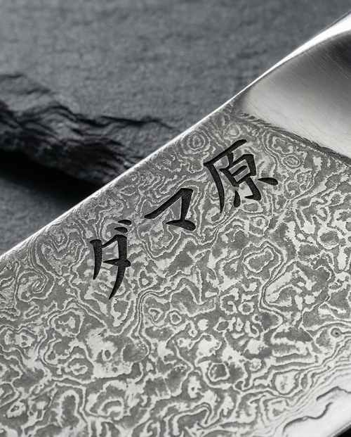 Gros plan sur lame en acier damassé d’un couteau japonais, motifs ondulés contrastés et gravure de caractères noirs.