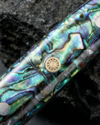 Gros plan sur manche de couteau japonais en résine nacrée bleu vert irisée, avec rivet métallique circulaire, posé sur pierre sombre.