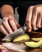 Couteau japonais Petty en acier damassé utilisé pour trancher un avocat sur planche en bois.