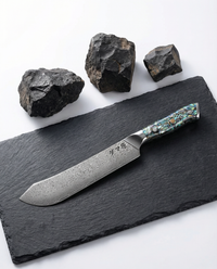 Couteau japonais Bunka en acier damassé posé sur ardoise noire, manche en résine nacrée bleu vert, pierres volcaniques en arrière-plan.