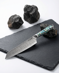 Couteau japonais kiritsuke en acier damassé posé sur ardoise noire, manche en résine bleu vert nacrée avec rivets métalliques, pierres volcaniques en fond.