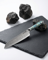 Couteau japonais kiritsuke en acier damassé posé sur ardoise noire, manche en résine bleu vert nacrée avec rivets métalliques, pierres volcaniques en fond.