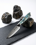 Couteau japonais Petty en acier damassé posé sur ardoise noire, manche en résine nacrée bleu vert avec rivets métalliques, pierres volcaniques en arrière-plan.
