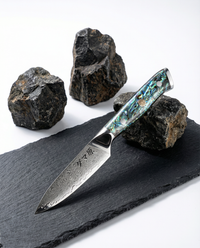 Couteau japonais Petty en acier damassé posé sur ardoise noire, manche en résine nacrée bleu vert avec rivets métalliques, pierres volcaniques en arrière-plan.