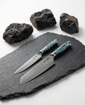 Deux couteaux japonais en acier damassé posés sur ardoise noire, manches en résine bleu vert nacrée, pierres volcaniques en arrière-plan.