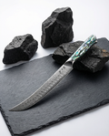 Couteau japonais Sujihiki en acier damassé posé sur ardoise noire, manche en résine bleu vert nacrée, pierres volcaniques en fond.