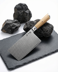 Couteau japonais nakiri en acier damassé, manche en bois clair, lame large rectangulaire sur ardoise noire avec roches volcaniques.
