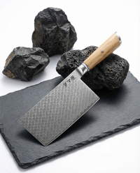Couteau japonais nakiri en acier damassé, manche en bois clair, lame large rectangulaire sur ardoise noire avec roches volcaniques.