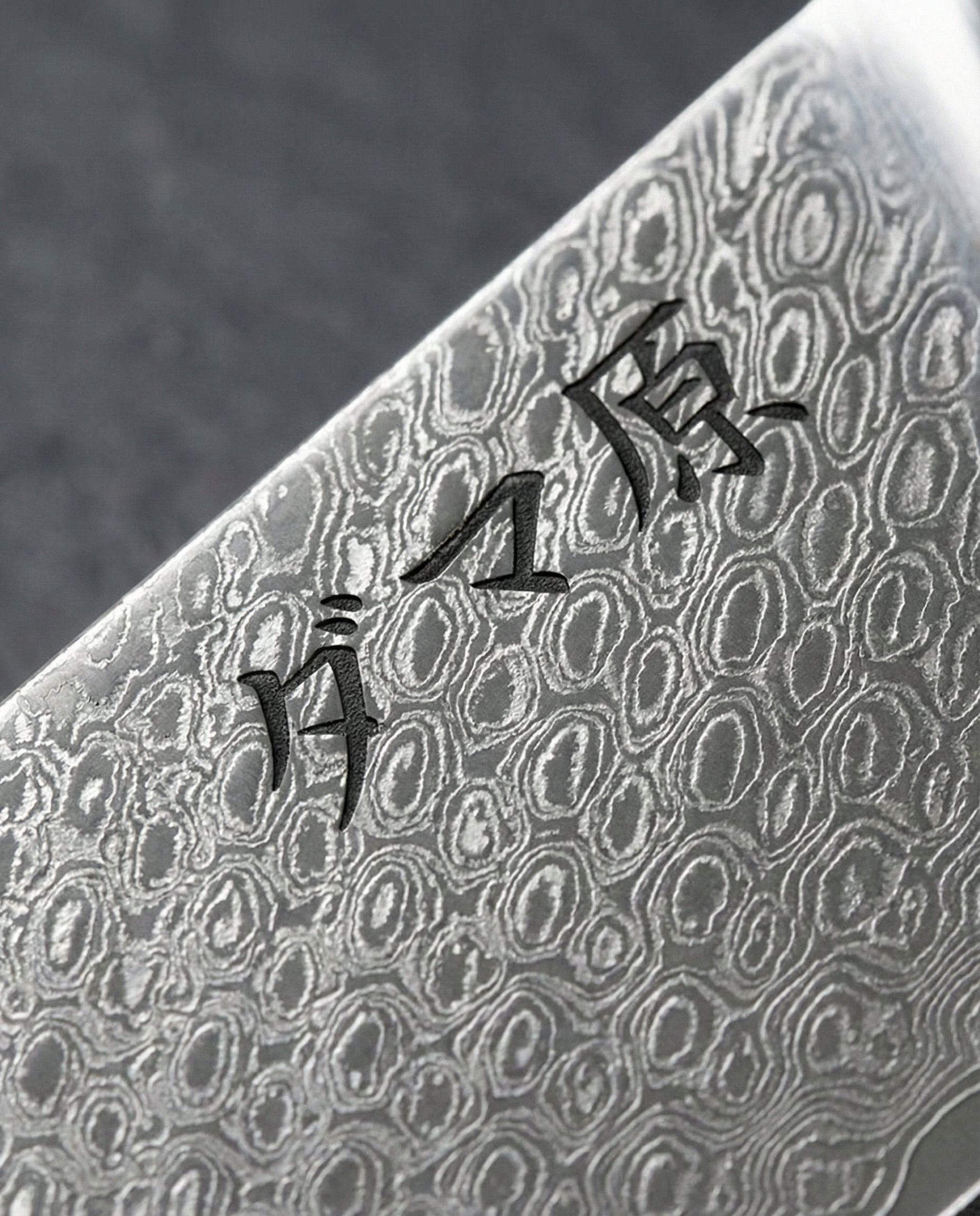 Gros plan sur lame de couteau japonais en acier damas, motif martelé circulaire et gravure damas origin noire près du talon.