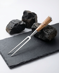 Fourchette à viande japonaise en acier inoxydable, manche en bois clair, posée sur ardoise noire avec roches brutes.