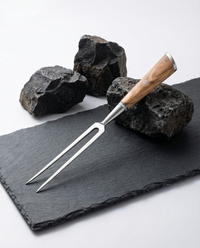 Fourchette à viande japonaise en acier inoxydable, manche en bois clair, posée sur ardoise noire avec roches brutes.