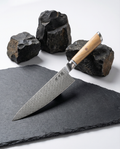 Couteau de chef japonais en acier damassé, manche en bois clair, présenté sur ardoise noire avec roches volcaniques