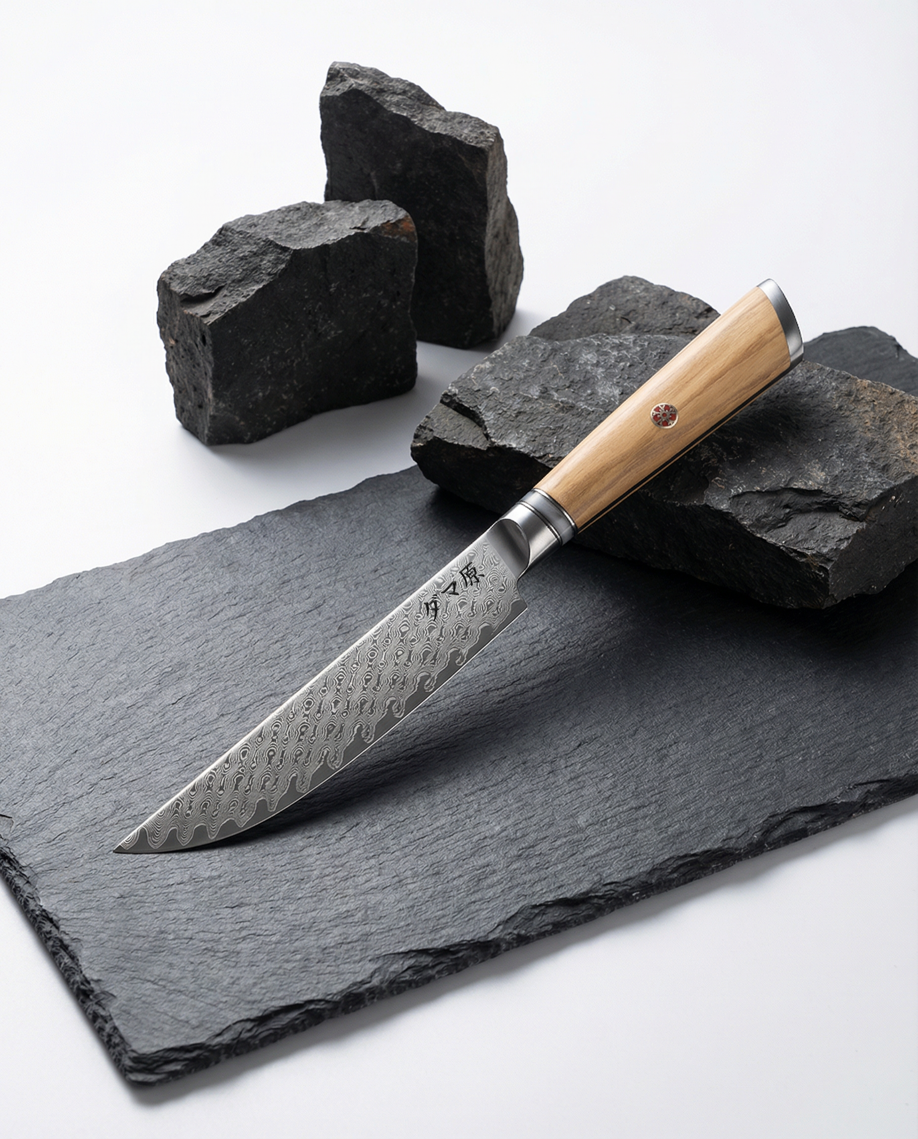 Couteau japonais petty en acier damassé, manche en bois clair, posé sur ardoise noire avec roches volcaniques décoratives.