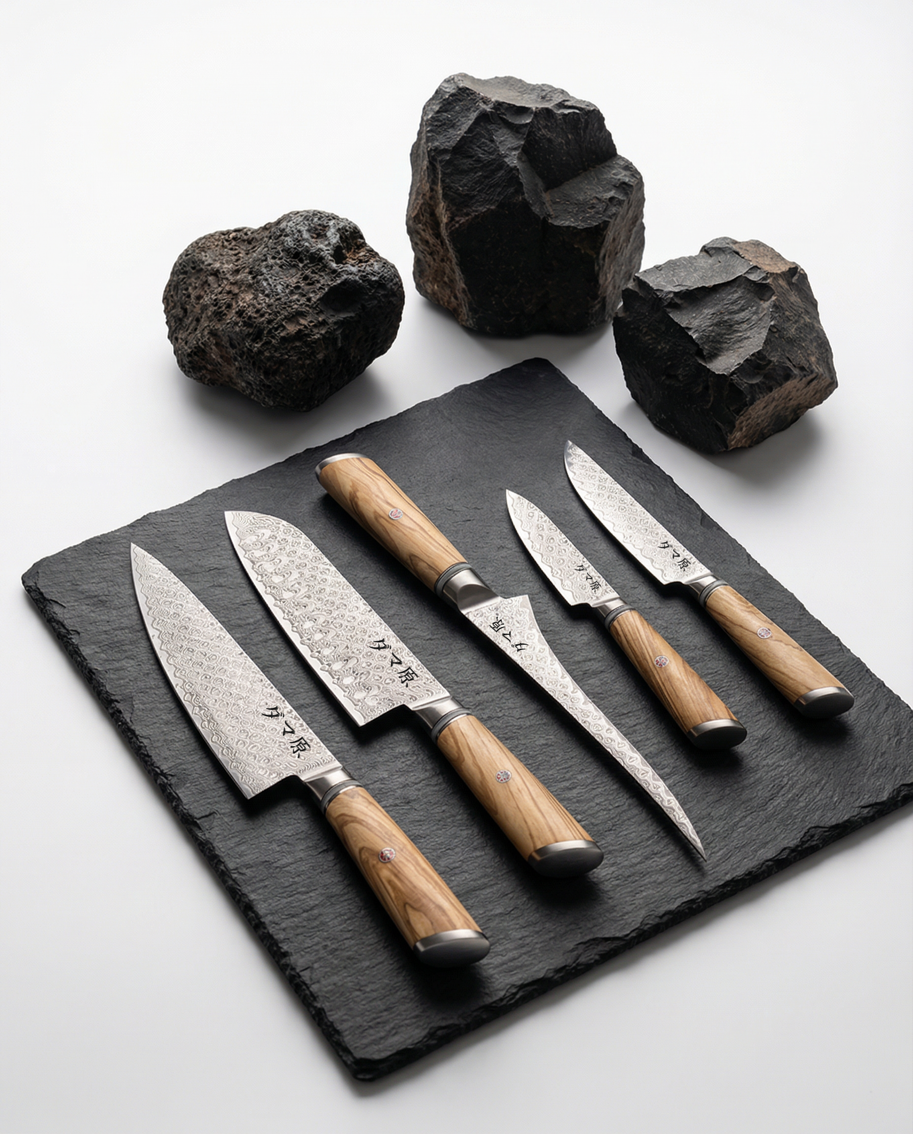 Set de 5 couteaux japonais en acier damas, manches bois clair, disposés sur plaque d’ardoise avec roches décoratives.