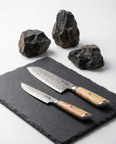 Set de 2 couteaux japonais en acier damas, manches bois clair naturel, posés sur ardoise noire avec roches volcaniques.