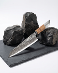 Couteau japonais en acier damassé à motif géométrique, manche en bois clair, posé sur ardoise noire avec pierres brutes