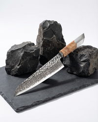 Couteau japonais en acier damassé à motif géométrique, manche en bois clair, posé sur ardoise noire avec pierres brutes