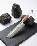 Couteau japonais en acier damassé à motif géométrique, manche en bois clair et résine transparente, posé sur ardoise noire avec pierres brutes