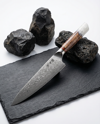 Couteau japonais chef en acier damas, manche bois et résine blanche, présenté sur ardoise noire avec pierres volcaniques.
