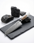 Couteau japonais chef en acier damas, manche bois ondé et résine blanche, posé sur ardoise noire avec pierres brutes.