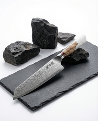 Couteau japonais chef en acier damas, manche bois ondé et résine blanche, posé sur ardoise noire avec pierres brutes.