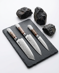 Set de 3 couteaux japonais damas avec manches bois et résine blanche, posés sur ardoise noire et pierres volcaniques.