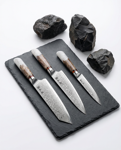 Set de 3 couteaux japonais damas avec manches bois et résine blanche, posés sur ardoise noire et pierres volcaniques.