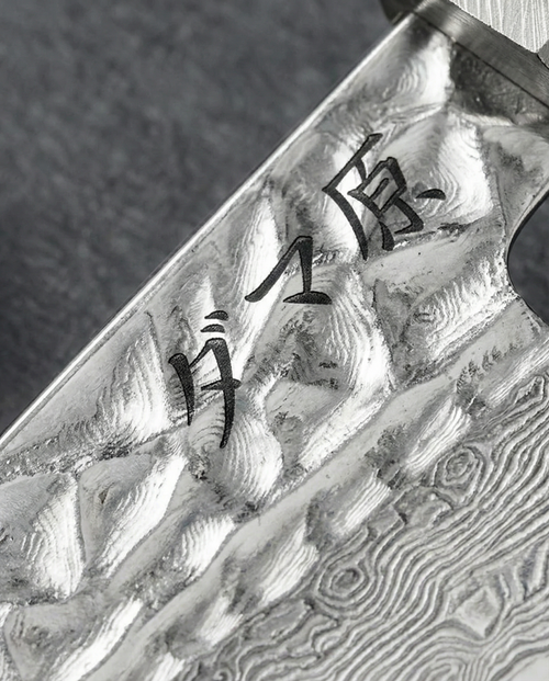 Gros plan sur la lame d’un couteau japonais en acier Damas 67 couches, avec motifs ondulés caractéristiques et gravure japonaise noire près du talon, finition martelée et texture brillante sur fond gris neutre.