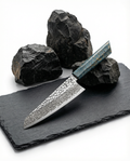 Couteau japonais Damas Origin avec lame en acier Damas 67 couches finition martelée et gravure japonaise, manche bleu azur marbré doré, posé sur une plaque d’ardoise noire avec pierres volcaniques en arrière-plan.