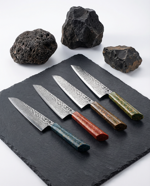 Pack de 4 couteaux japonais Damas Origin avec lames en acier Damas 67 couches et manches colorés (bleu azur, rouge volcan, brun doré et vert jungle) disposés sur une plaque d’ardoise noire, avec pierres volcaniques en arrière-plan.