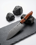 Couteau japonais santoku en acier damas, manche ergonomique en bois rouge, posé sur ardoise avec pierres noires.