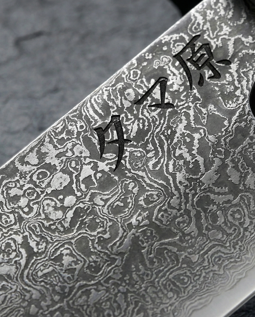 Gros plan sur lame de couteau japonais en acier damassé, motifs ondulés contrastés et gravure de caractères japonais noirs.